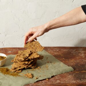 A hand grabs a cracker from a stack of chickpea seed crackers that sit on a piece of parchment paper on a wooden table.