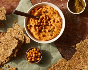 A white bowl is filled with harissa carrot hummus. The hummus is garnished with chickpeas and a stack of seed crackers sits on the side of the bowl.