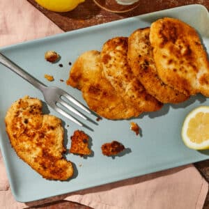 Almond Crusted Chicken Cutlets on a matte slate blue platter with a lemon wedge and pewter serving fork on a salmon colored napkin.