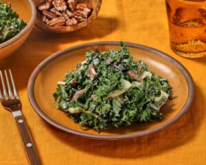 Kale salad with parmesan dressing on a mustard colored plate on a mustard colored linen tablecloth with a fork, glass of water and bowl of toasted pecans.
