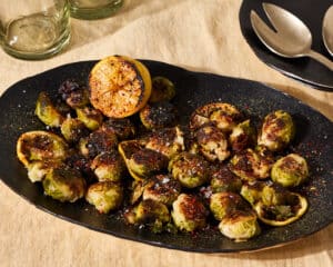 Crispy Smashed Brussels Sprouts in. a black oval platter with a charred lemon on a light yellow linen tablecloth.