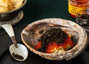 Caviar Pie topped with black osetra caviar and bright orange trout roe, presented in a large abalone shell with a mother-of-pearl spoon and kettle chips in the background on a green velvet cloth.