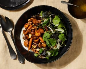 Roasted Root Veggie Salad with EAsy Lemon Dressing in a black salad bowl on a butter colored tablecloth with a black spoon and fork and a couple of side plates peeking in.