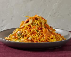 Apple & Beet Slaw with Carrot Ginger Dressing in a dark metallic glazed shallow bowl on a burgundy linen tablecloth with a white plaster wall in the background.