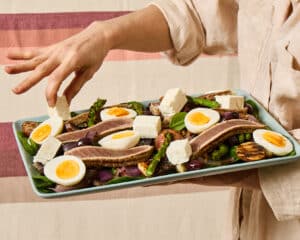 Spring Nicoise Salad on a light blue platter. Hand reaching above it against a background of textured linen cloth in cream, dusty rose and dusty pink stripes.