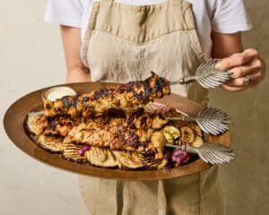 Berbere Chicken Skewers on a bed of grilled vegetables on a copper platter being held by a woman wearing a tan linen apron and white tshirt.
