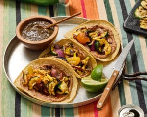 3 Vegan Charred Veggie & Refried Bean Tacos on an aluminum grill pan with a wedged lime and a small wood bowl with Yucatecan salsa and wood spoon. Wood handled knife resting on pan. Blue/green/orang/red striped linen tablecloth and a can of soda water in the right bottom corner.