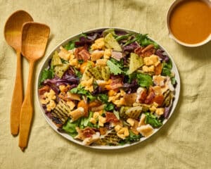 Smoked Trout and Green Tomato Salad on an oval platter on a light green linen cloth. Small bowl of roasted tomato dressing in top right corner, olive wood serving utensils on left side of platter.