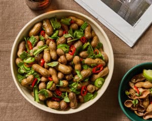 Vietnamese boiled peanuts in a white bowl with fresh herbs and red and green chilies. On a brown linen tablecloth. Amber glass and newspaper peaking in at the top, Small green bowl with discarded peanut shells in bottom right.