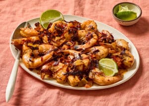 Guajillo Garlic Shrimp on an oval white platter with a white serving spoon tucked in. A bowl of cut limes is in the top right corner. Platter sits on a dusty rose linen tablecloth.