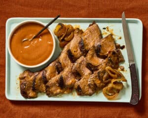 Georgia Peach & Vidalia BBQ Sauce in a small white bowl on a light blue platter with grilled pork. A wood handled knife rests on the right side of the platter as it sits on a heavy burnt orange linen cloth.