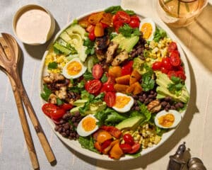 Southwestern Cobb with Creamy Chipotle Dressing on a round white platter on a cream and light blue textile. Wooden serving spoons sit to the left of the plate. A small bowl of dressing is on the top left, small glass pitcher on the top right. Antique salt and pepper shakers on the bottom right.