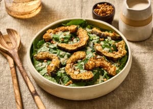Kale salad with Parmesan crusted delicata squash croutons, toasted pine nuts and shaved Parmesan in a shallow ivory ceramic bowl on a natural linen surface. Olive wood salad servers rest to the left, with a small bowl of pine nuts and a ceramic pitcher of creamy Parmesan dressing behind the salad.