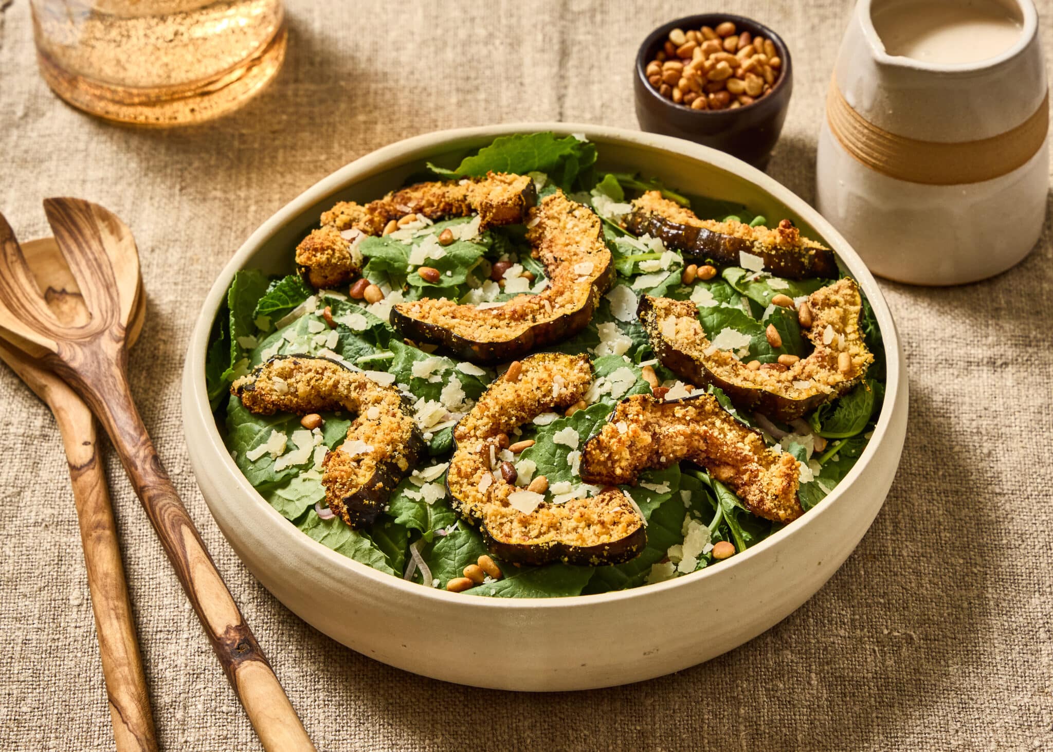 Kale salad with Parmesan crusted delicata squash croutons, toasted pine nuts and shaved Parmesan in a shallow ivory ceramic bowl on a natural linen surface. Olive wood salad servers rest to the left, with a small bowl of pine nuts and a ceramic pitcher of creamy Parmesan dressing behind the salad.