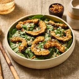 Kale salad with Parmesan crusted delicata squash croutons, toasted pine nuts and shaved Parmesan in a shallow ivory ceramic bowl on a natural linen surface. Olive wood salad servers rest to the left, with a small bowl of pine nuts and a ceramic pitcher of creamy Parmesan dressing behind the salad.