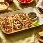 Three primal blend chicken tacos on a white enamel tray with green trim on a light green linen tablecloth. Small wooden bowls are scattered around with cheese, tomatoes, avocado, limes, corn tortillas and there's also a bottle of Valentina hot sauce.