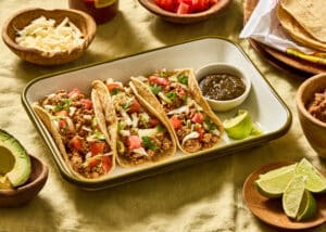 Three primal blend chicken tacos on a white enamel tray with green trim on a light green linen tablecloth. Small wooden bowls are scattered around with cheese, tomatoes, avocado, limes, corn tortillas and there's also a bottle of Valentina hot sauce.